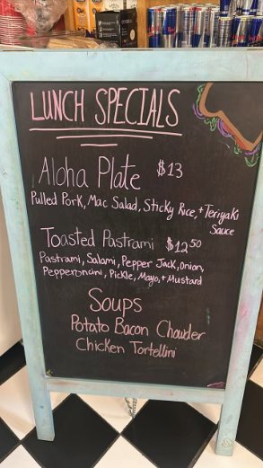 A blue A-frame chalkboard inside the cafe, hand lettered in coloured chalk. It reads Lunch Specials, Aloha Plate thirteen dollars, Toasted Pastrami twelve fifty, and Soups, potato bacon chowder and chicken tortellini.